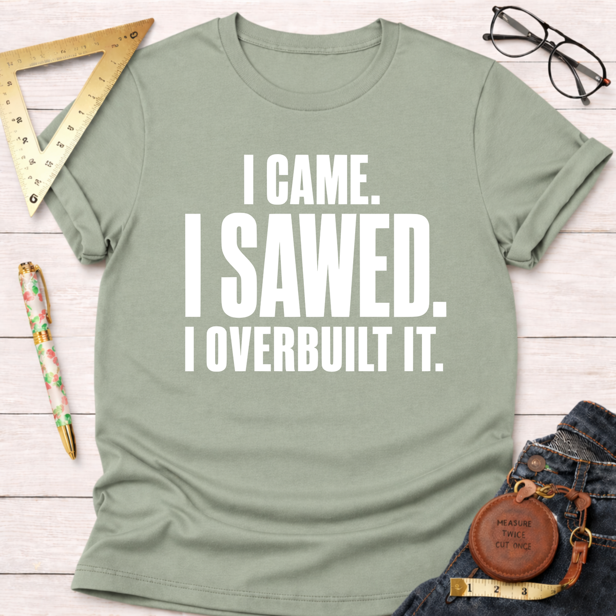 Sage green t-shirt with "I CAME. I SAWED. I OVERBUILT IT." in white text, styled with jeans and tools.