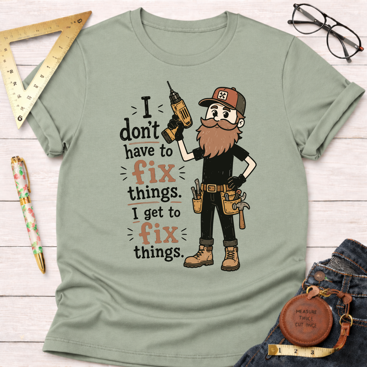 Sage green cotton t-shirt with a cartoon carpenter holding a drill and the text "I don't have to fix things. I get to fix things.