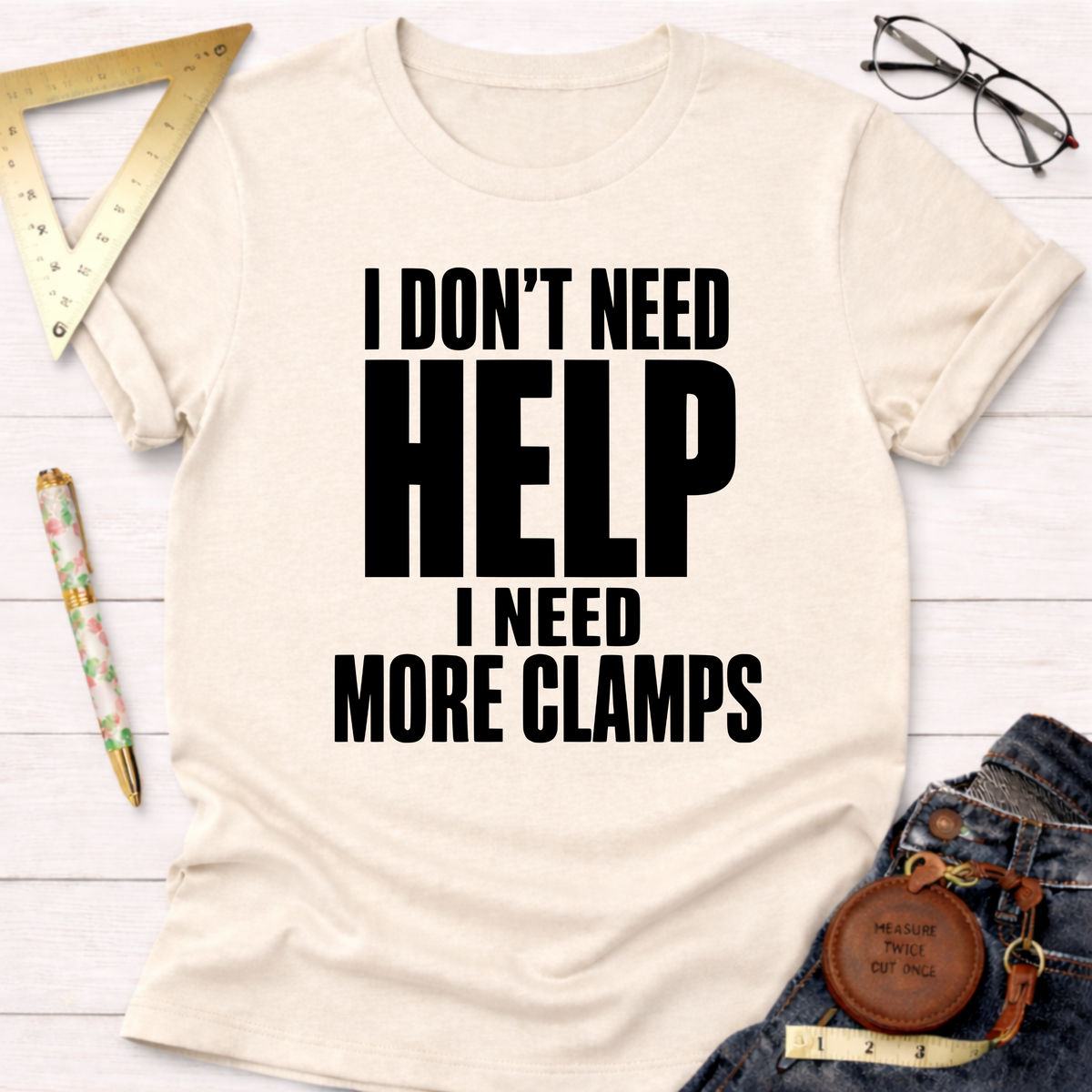 Unisex cream cotton t-shirt with "I DON'T NEED HELP. I NEED MORE CLAMPS" in bold black text.