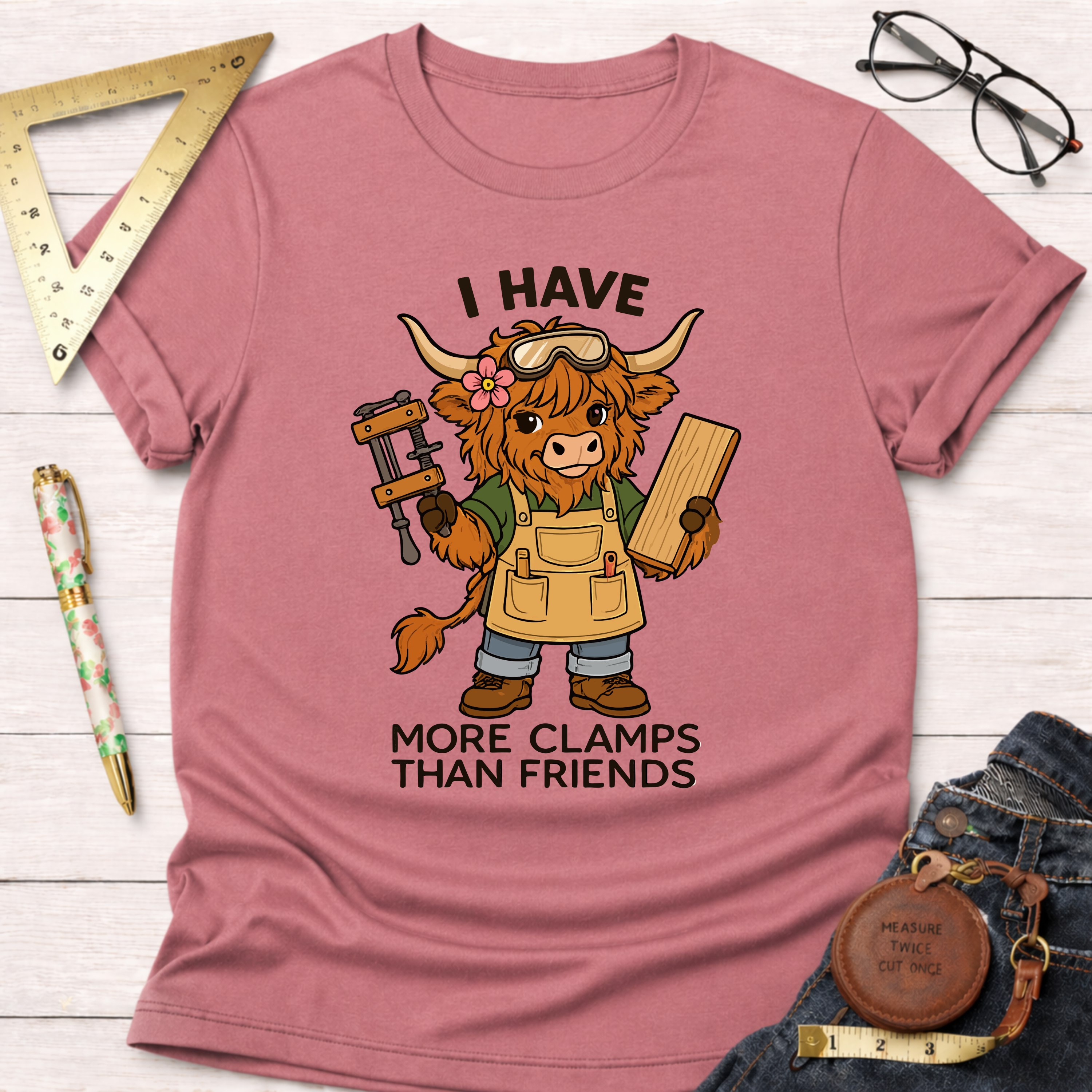 Women's funny woodworking graphic tee shirt for female woodworkers, makers, and DIY builders — Sawdust & Ponytails. Dusty rose t-shirt with a cartoon Highland cow wearing safety goggles and an apron, holding a clamp and wood, with text "I HAVE MORE CLAMPS THAN FRIENDS.