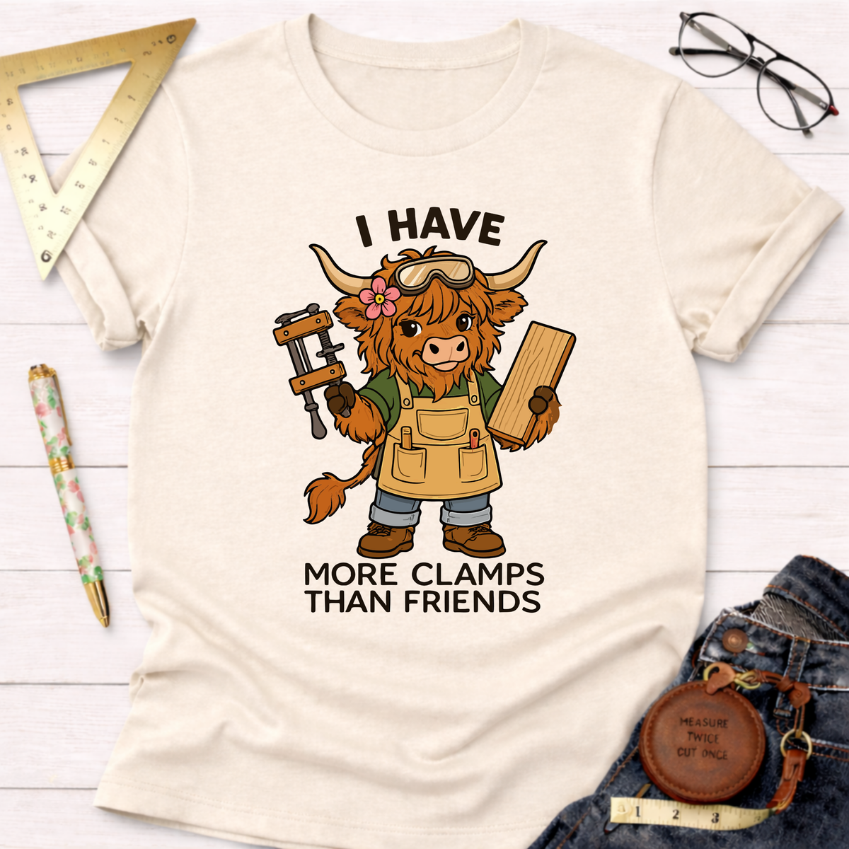 Women's funny woodworking graphic tee shirt for female woodworkers, makers, and DIY builders — Sawdust & Ponytails. Cream t-shirt with a cartoon Highland cow wearing goggles and an apron, holding a clamp and wood, with text "I Have More Clamps Than Friends".