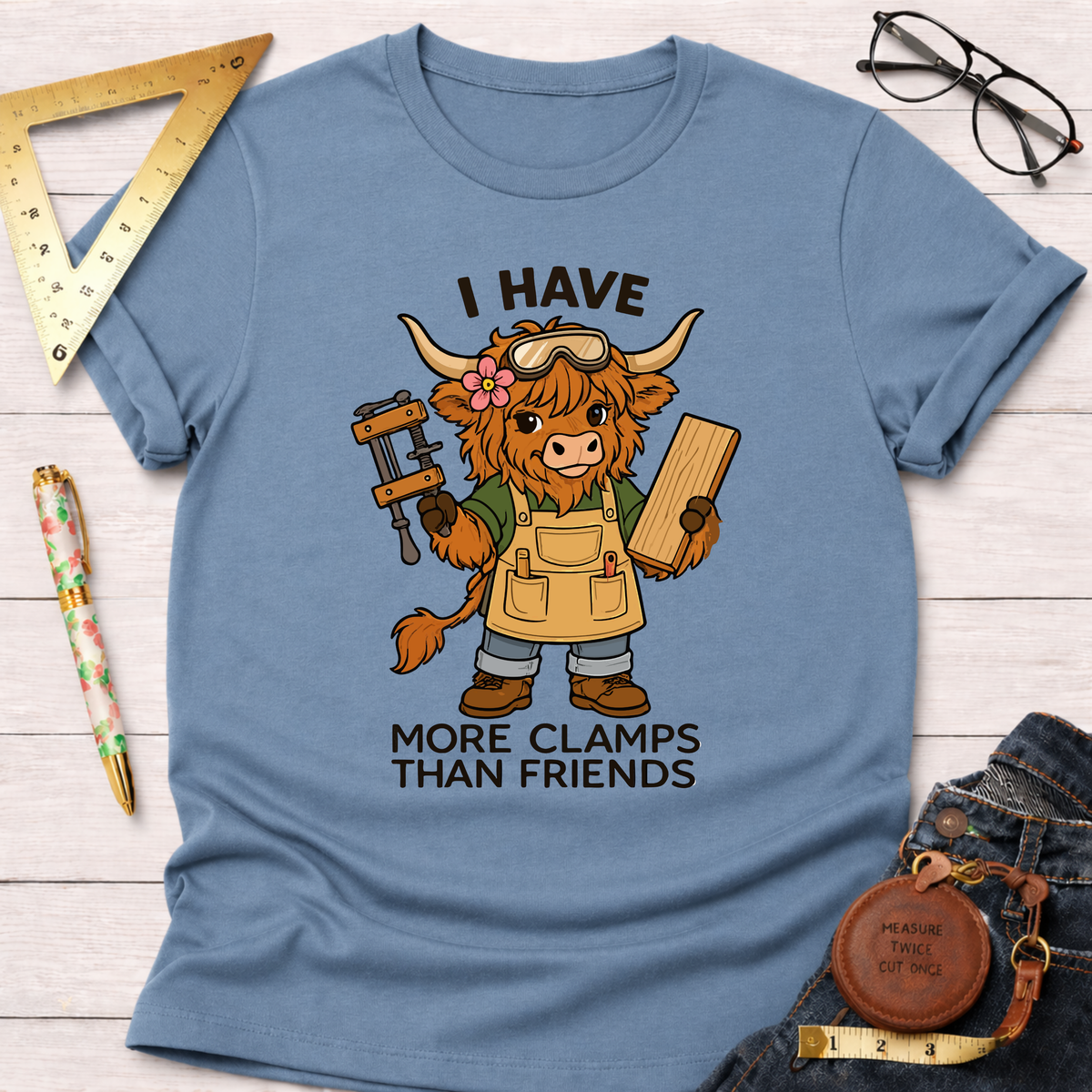 Women's funny woodworking graphic tee shirt for female woodworkers, makers, and DIY builders — Sawdust & Ponytails. Light blue cotton tee with a cartoon highland cow wearing goggles and an apron, holding a clamp and wood, with text "I Have More Clamps Than Friends".
