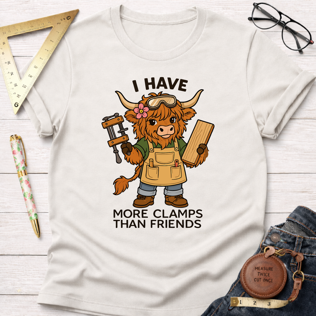 Women's funny woodworking graphic tee shirt for female woodworkers, makers, and DIY builders — Sawdust & Ponytails. White cotton tee with a cartoon Highland cow wearing goggles, an apron, holding a clamp and wood, with the text "I HAVE MORE CLAMPS THAN FRIENDS".
