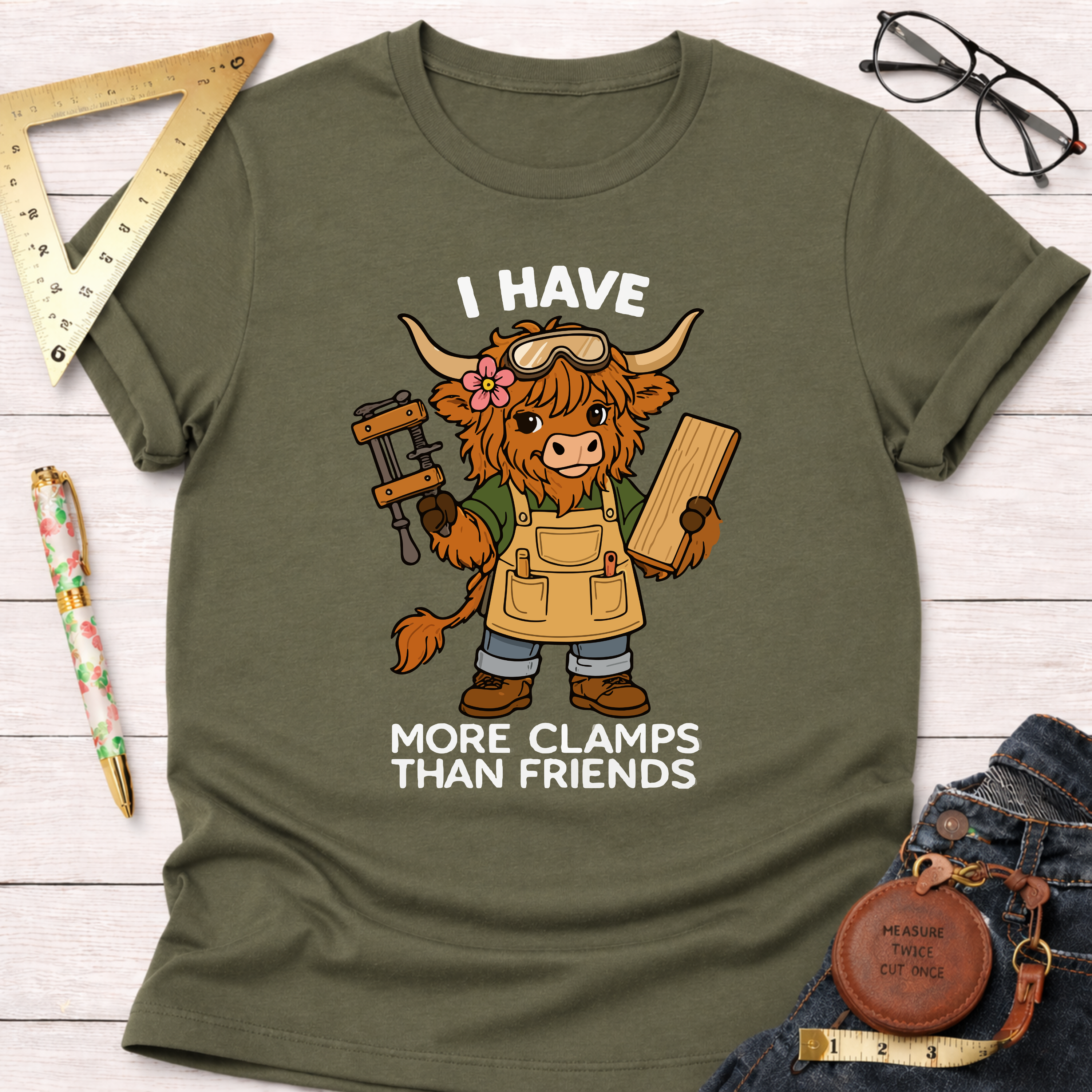Women's funny woodworking graphic tee shirt for female woodworkers, makers, and DIY builders — Sawdust & Ponytails. Olive green cotton tee with a cartoon Highland cow in an apron, holding a clamp and wood, with "I Have More Clamps Than Friends" text.
