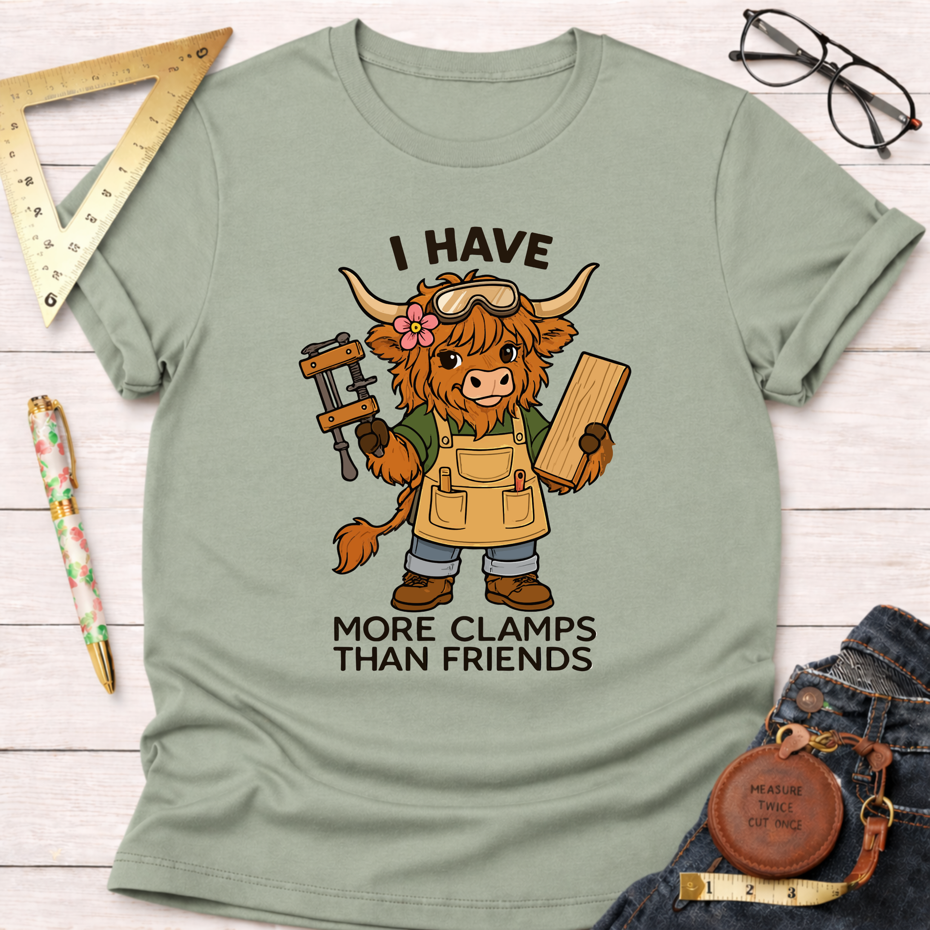 Women's funny woodworking graphic tee shirt for female woodworkers, makers, and DIY builders — Sawdust & Ponytails. Sage green t-shirt with a cartoon Highland cow wearing safety goggles and an apron, holding a clamp and wood, with text "I Have More Clamps Than Friends.