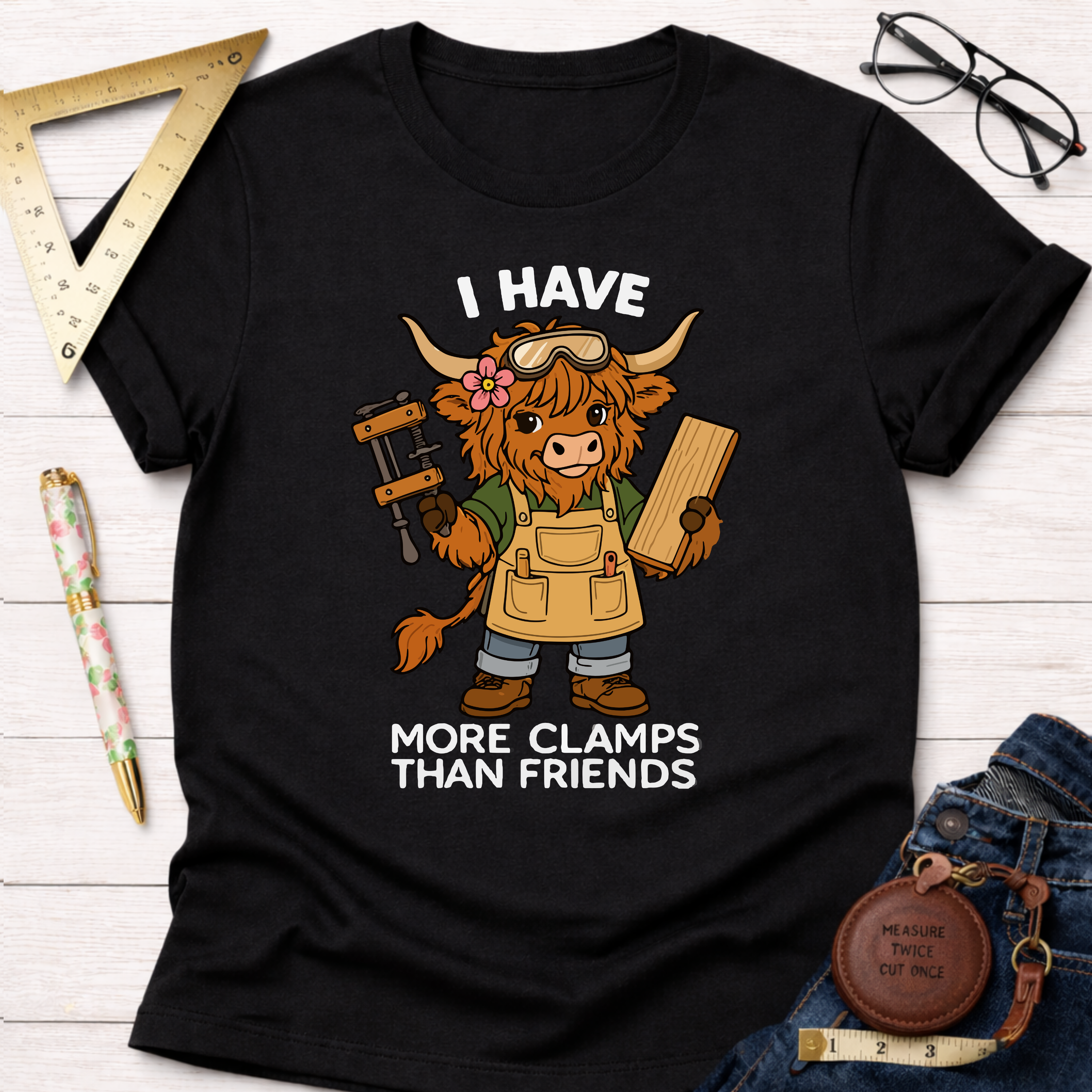 Women's funny woodworking graphic tee shirt for female woodworkers, makers, and DIY builders — Sawdust & Ponytails. Black cotton tee with a cartoon Highland cow wearing safety glasses and an apron, holding a clamp and wood.
