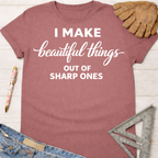 Women's funny woodworking graphic tee shirt for female woodworkers, makers, and DIY builders — Sawdust & Ponytails. Mauve cotton t-shirt with "I Make beautiful things OUT OF SHARP ONES" in white text, styled with jeans, a pen, a leather glove, and a metal square ruler.