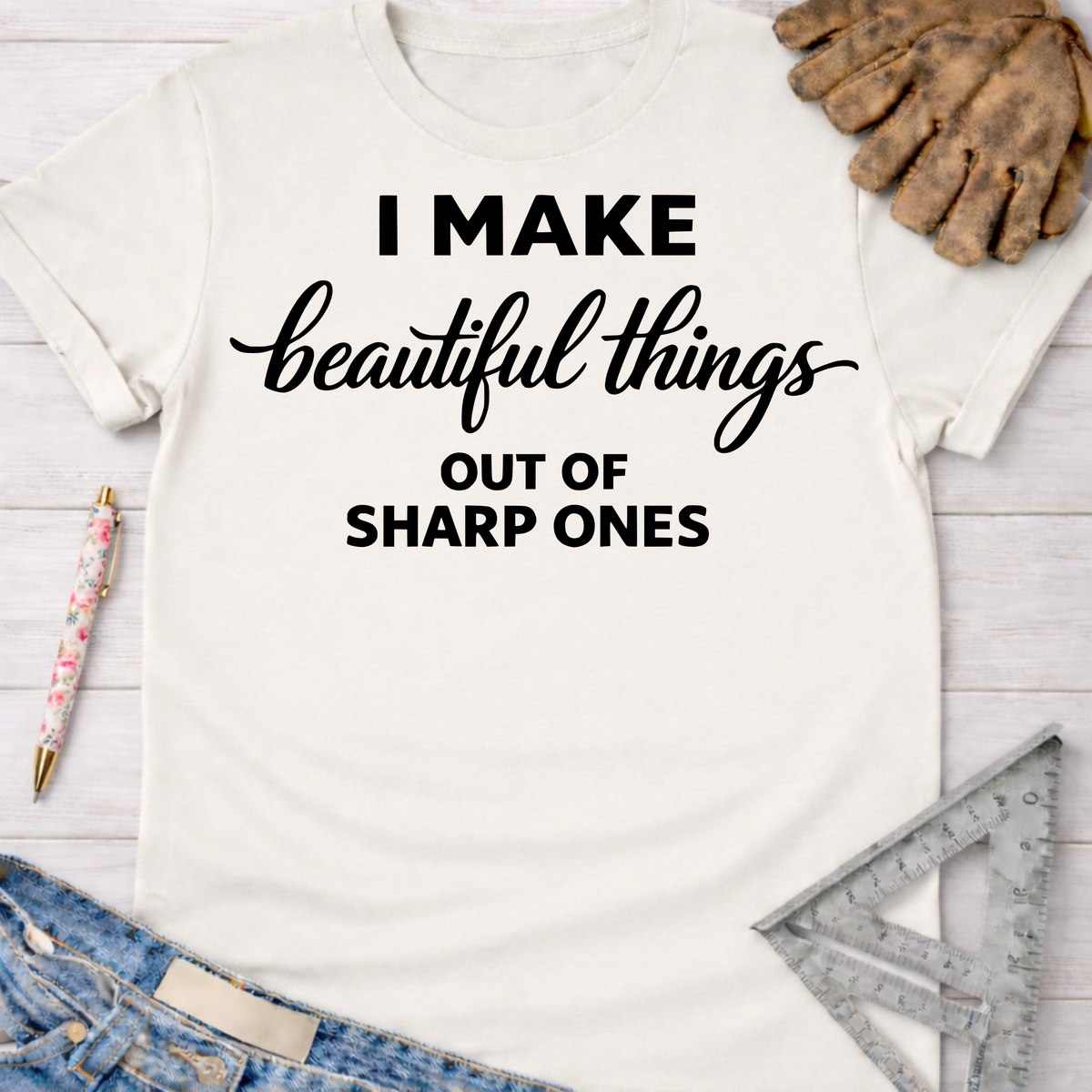 Women's funny woodworking graphic tee shirt for female woodworkers, makers, and DIY builders — Sawdust & Ponytails. Cream t-shirt with "I MAKE beautiful things OUT OF SHARP ONES" text, shown with jeans, work gloves, and tools.