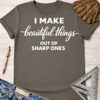 Women's funny woodworking graphic tee shirt for female woodworkers, makers, and DIY builders — Sawdust & Ponytails. Dark gray cotton tee with "I Make beautiful things OUT OF SHARP ONES" in white text, styled with jeans, gloves, and tools.