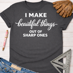 Charcoal gray women's "I Make Beautiful Things Out Of Sharp Ones" graphic tee with work glove, jeans, and ruler.