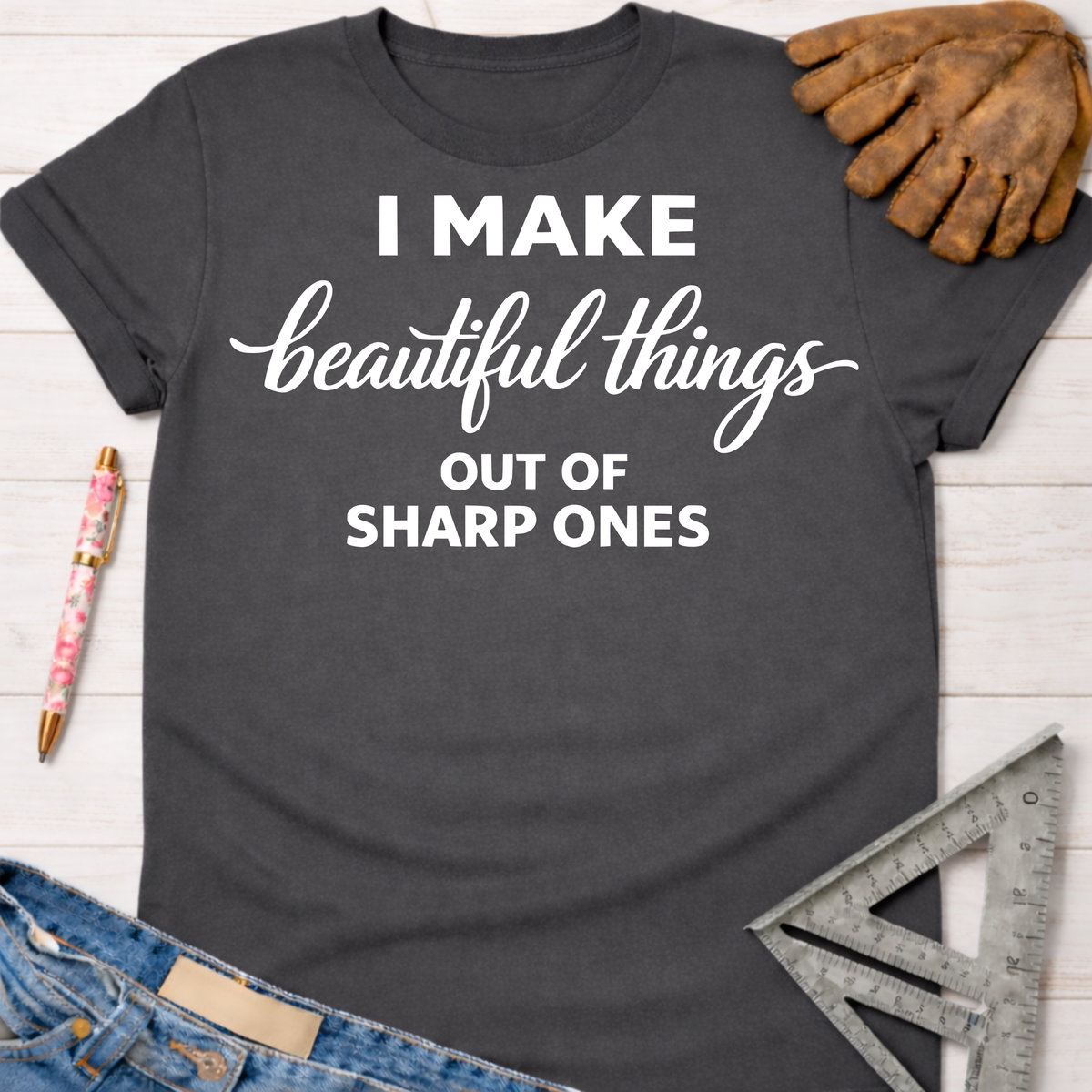 Charcoal gray women's "I Make Beautiful Things Out Of Sharp Ones" graphic tee with work glove, jeans, and ruler.