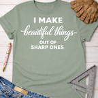 Women's funny woodworking graphic tee shirt for female woodworkers, makers, and DIY builders — Sawdust & Ponytails. Sage green cotton tee with "I Make beautiful things OUT OF SHARP ONES" in white text.