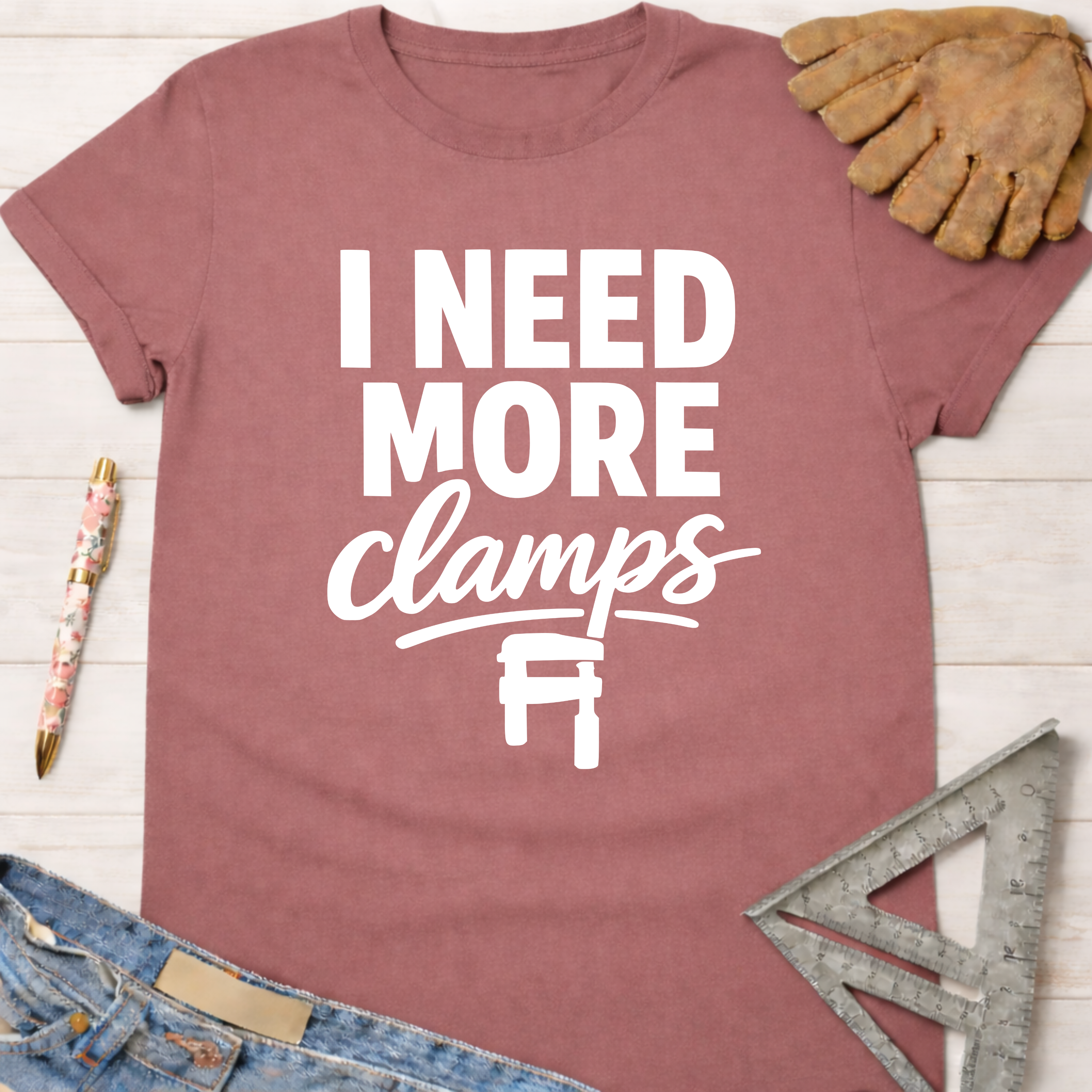 Women's funny woodworking graphic tee shirt for female woodworkers, makers, and DIY builders — Sawdust & Ponytails. Mauve short-sleeve t-shirt with "I NEED MORE clamps" text and clamp graphic.