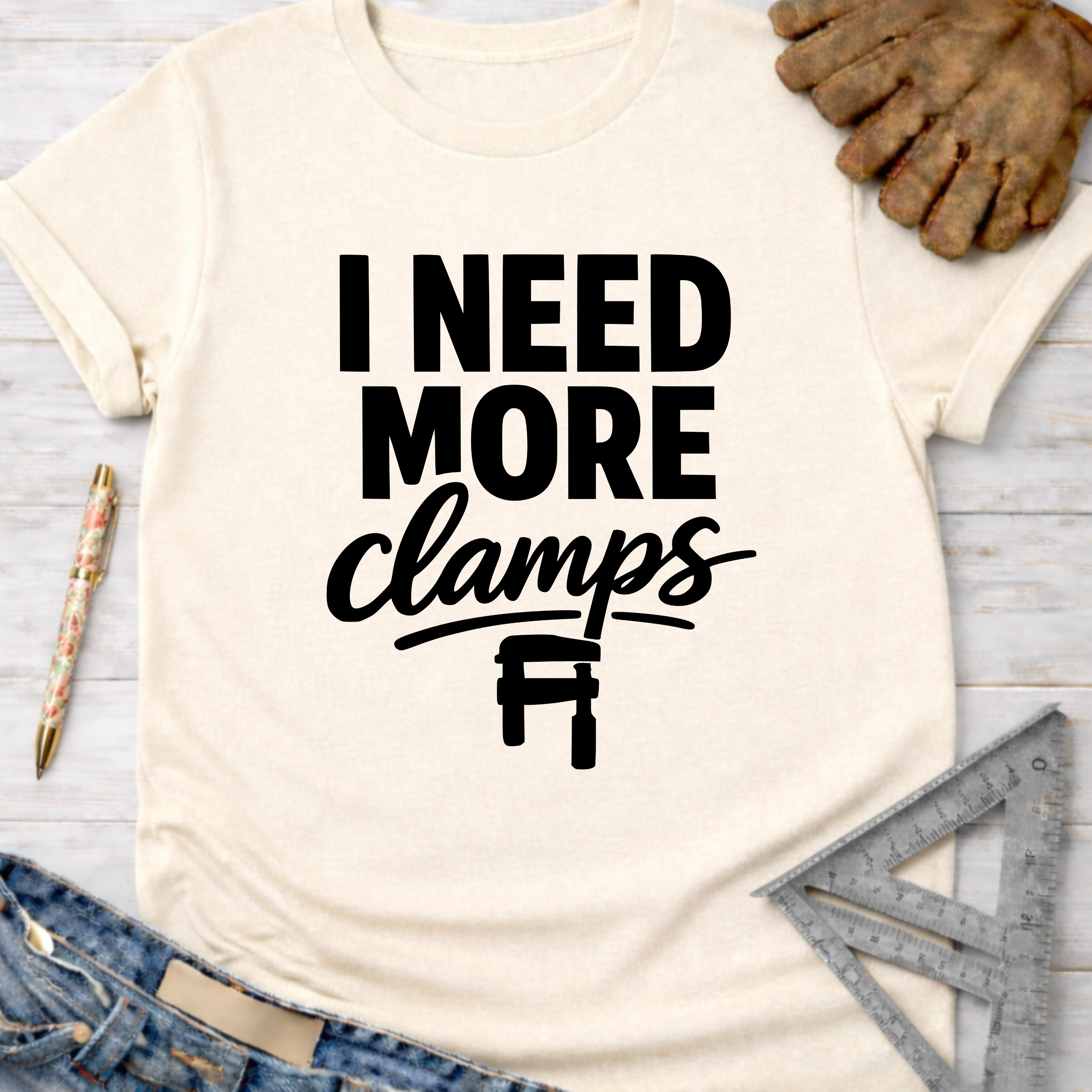 Women's funny woodworking graphic tee shirt for female woodworkers, makers, and DIY builders — Sawdust & Ponytails. Cream t-shirt with "I Need More Clamps" text and clamp graphic.