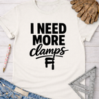 Women's funny woodworking graphic tee shirt for female woodworkers, makers, and DIY builders — Sawdust & Ponytails. White cotton t-shirt with "I NEED MORE clamps" in black text and a clamp graphic.