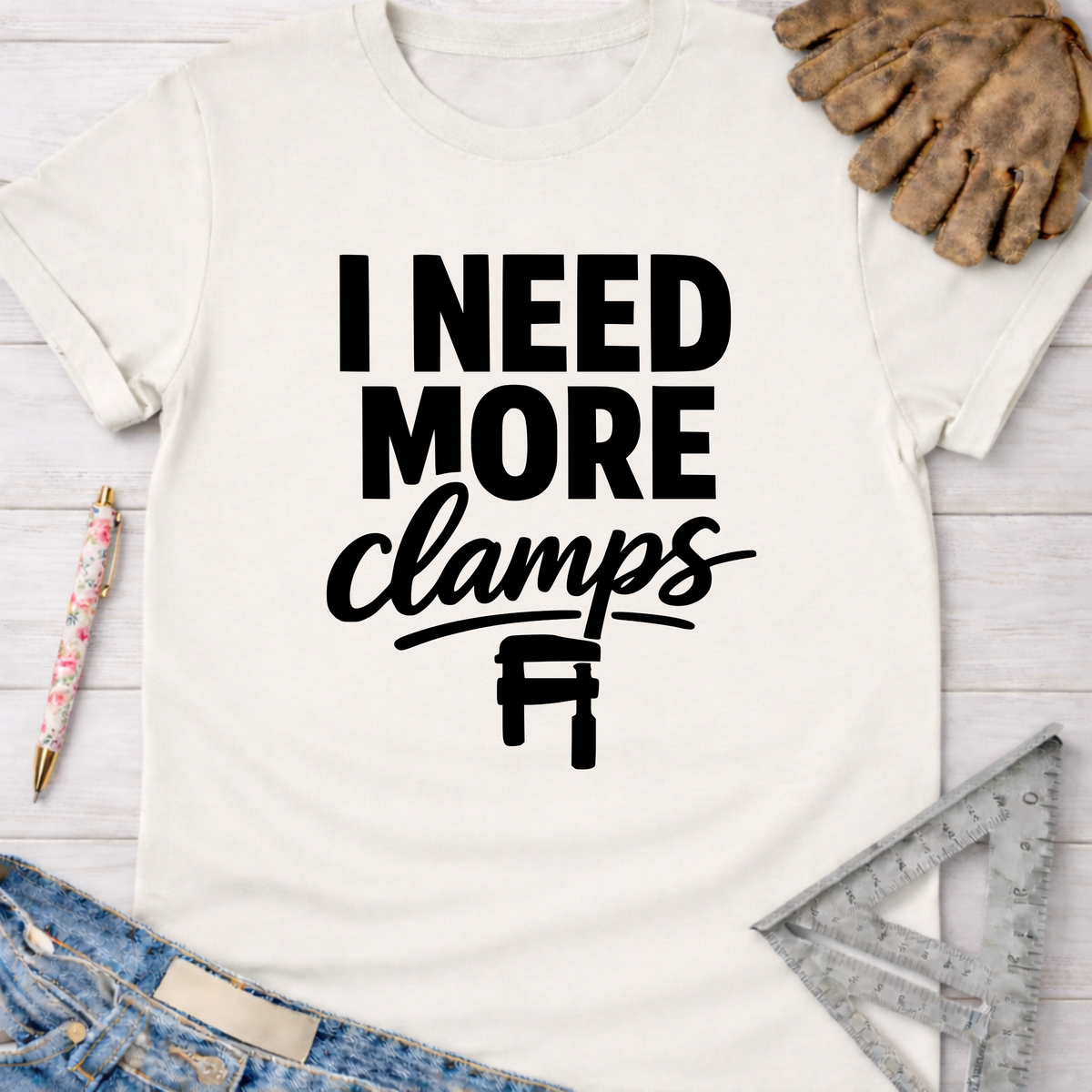 Women's funny woodworking graphic tee shirt for female woodworkers, makers, and DIY builders — Sawdust & Ponytails. White cotton t-shirt with "I NEED MORE clamps" in black text and a clamp graphic.