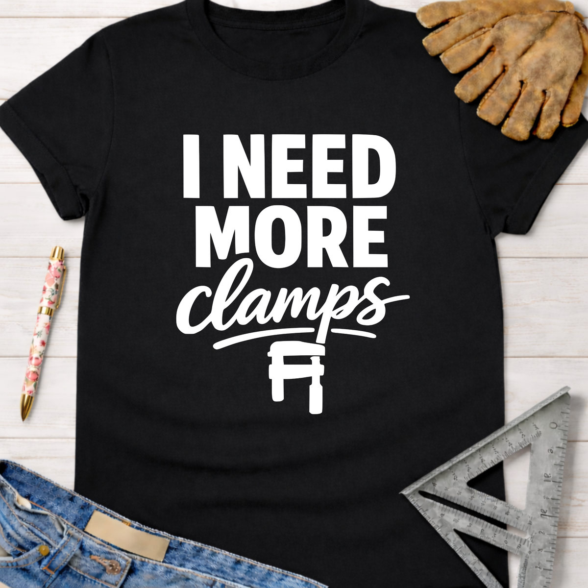 Women's funny woodworking graphic tee shirt for female woodworkers, makers, and DIY builders — Sawdust & Ponytails. Black cotton t-shirt with "I NEED MORE clamps" in white text and a clamp icon.