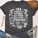Women's funny woodworking graphic tee shirt for female woodworkers, makers, and DIY builders — Sawdust & Ponytails. Dark gray cotton t-shirt with "YES I REALLY DO NEED ALL THESE Power Tools" in white, surrounded by various power tool illustrations.