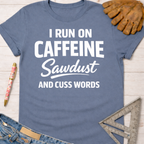 Women's funny woodworking graphic tee shirt for female woodworkers, makers, and DIY builders — Sawdust & Ponytails. Blue cotton tee with "I RUN ON CAFFEINE Sawdust AND CUSS WORDS" in white text.