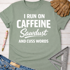 Women's funny woodworking graphic tee shirt for female woodworkers, makers, and DIY builders — Sawdust & Ponytails. Sage green cotton t-shirt with "I RUN ON CAFFEINE Sawdust AND CUSS WORDS" in white text.