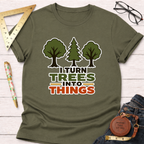 Olive green "I Turn Trees Into Things" t-shirt with tree graphics, styled on a white wooden surface.