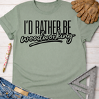 Women's funny woodworking graphic tee shirt for female woodworkers, makers, and DIY builders — Sawdust & Ponytails. Sage green cotton tee with "I'd Rather Be Woodworking" in black text.