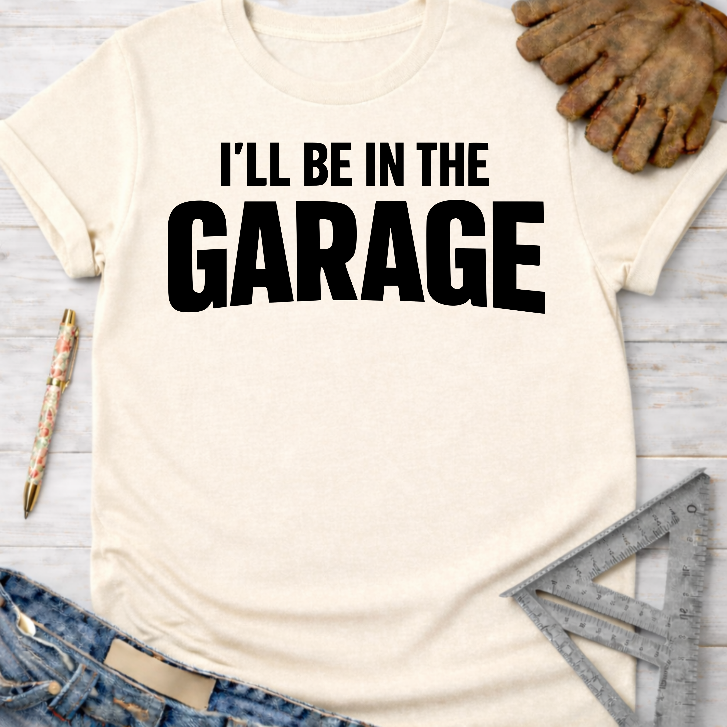 Women's funny woodworking graphic tee shirt for female woodworkers, makers, and DIY builders — Sawdust & Ponytails. Cream t-shirt with "I'll Be In The GARAGE" in black text, styled with jeans, work gloves, and a ruler.