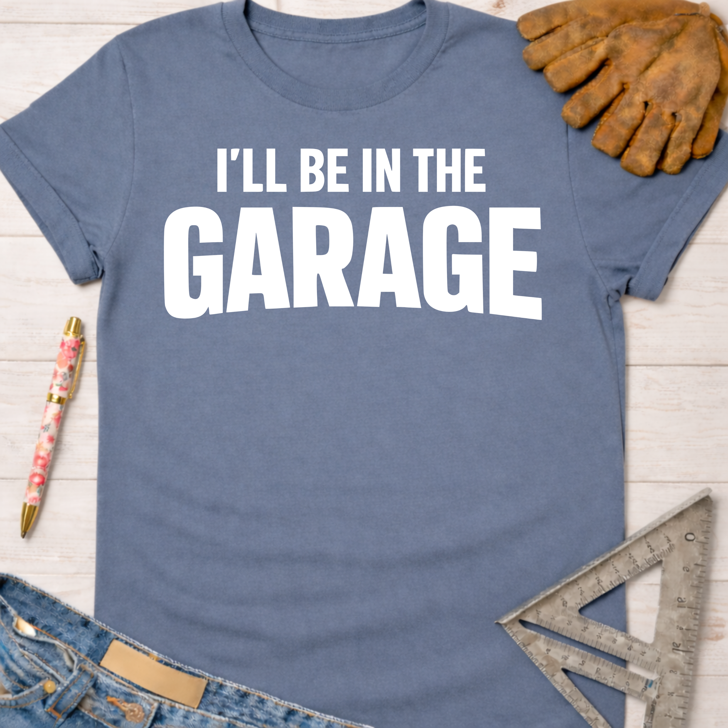 Women's funny woodworking graphic tee shirt for female woodworkers, makers, and DIY builders — Sawdust & Ponytails. Blue cotton t-shirt with "I'LL BE IN THE GARAGE" in white text.