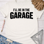 Women's funny woodworking graphic tee shirt for female woodworkers, makers, and DIY builders — Sawdust & Ponytails. White cotton tee with "I'LL BE IN THE GARAGE" in black text.