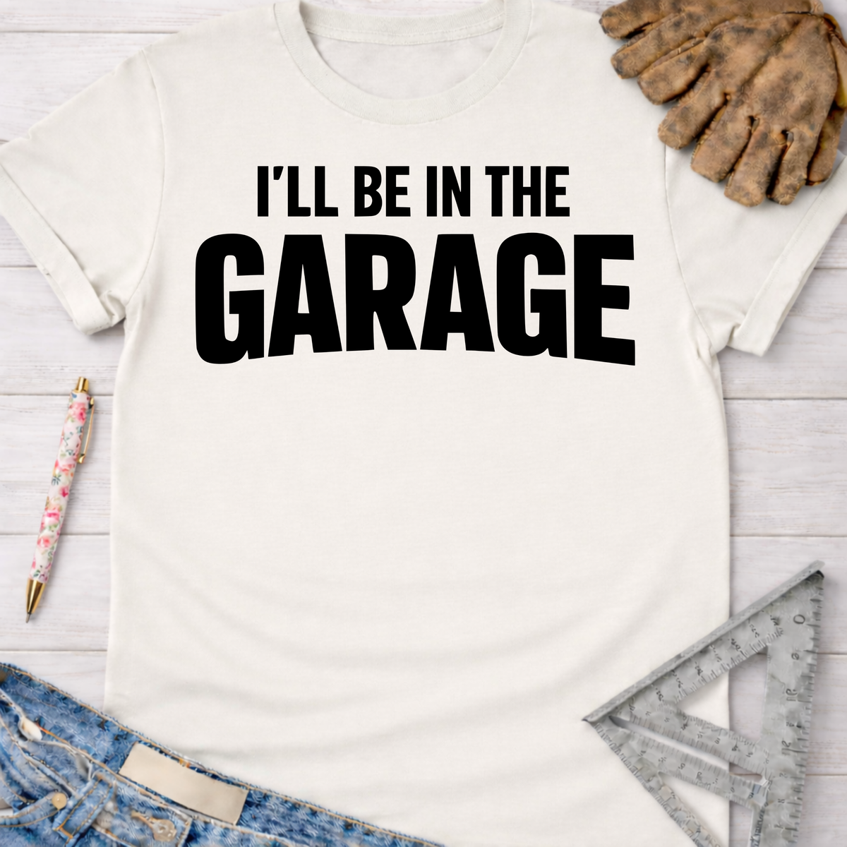 Women's funny woodworking graphic tee shirt for female woodworkers, makers, and DIY builders — Sawdust & Ponytails. White cotton tee with "I'LL BE IN THE GARAGE" in black text.