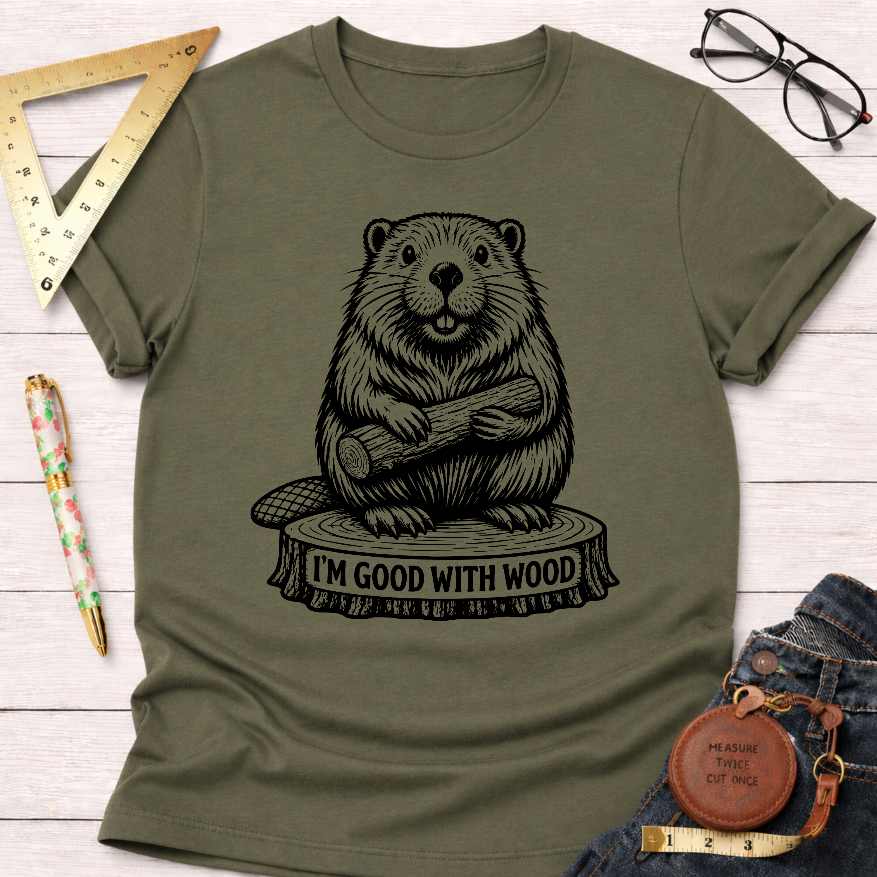 Women's funny woodworking graphic tee shirt for female woodworkers, makers, and DIY builders — Sawdust & Ponytails. Olive green cotton tee with a black beaver holding wood on a tree stump, "I'M GOOD WITH WOOD" text.