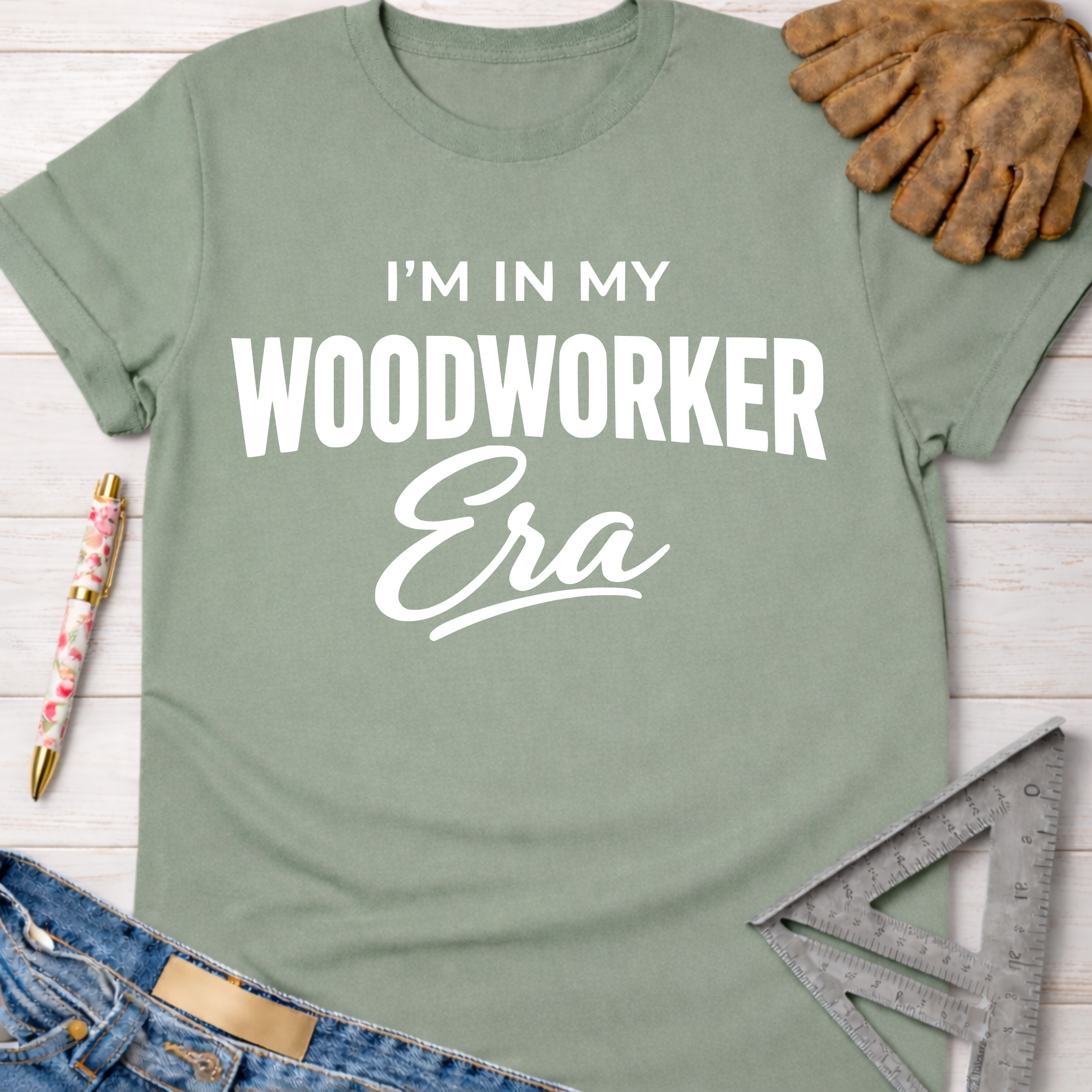 Women's funny woodworking graphic tee shirt for female woodworkers, makers, and DIY builders — Sawdust & Ponytails. Sage green cotton tee with "I'm In My Woodworker Era" in white text, next to work gloves and tools.