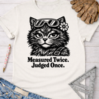 Women's funny woodworking graphic tee shirt for female woodworkers, makers, and DIY builders — Sawdust & Ponytails. Cream t-shirt with a black graphic of a cat wearing safety goggles and a flower, with "Measured Twice. Judged Once." text.