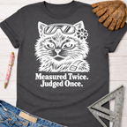 Women's funny woodworking graphic tee shirt for female woodworkers, makers, and DIY builders — Sawdust & Ponytails. Dark gray tee with white "Judgy Gato" cat wearing safety glasses and flower, text "Measured Twice. Judged Once.