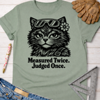 Women's funny woodworking graphic tee shirt for female woodworkers, makers, and DIY builders — Sawdust & Ponytails. Sage green cotton tee with a black graphic of a cat in safety goggles and a flower, text reads "Measured Twice. Judged Once.