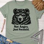 Women's funny woodworking graphic tee shirt for female woodworkers, makers, and DIY builders — Sawdust & Ponytails. Sage green cotton t-shirt with a black graphic of a focused bear wearing goggles and a flower, with the text "Not Angry. Just Focused.