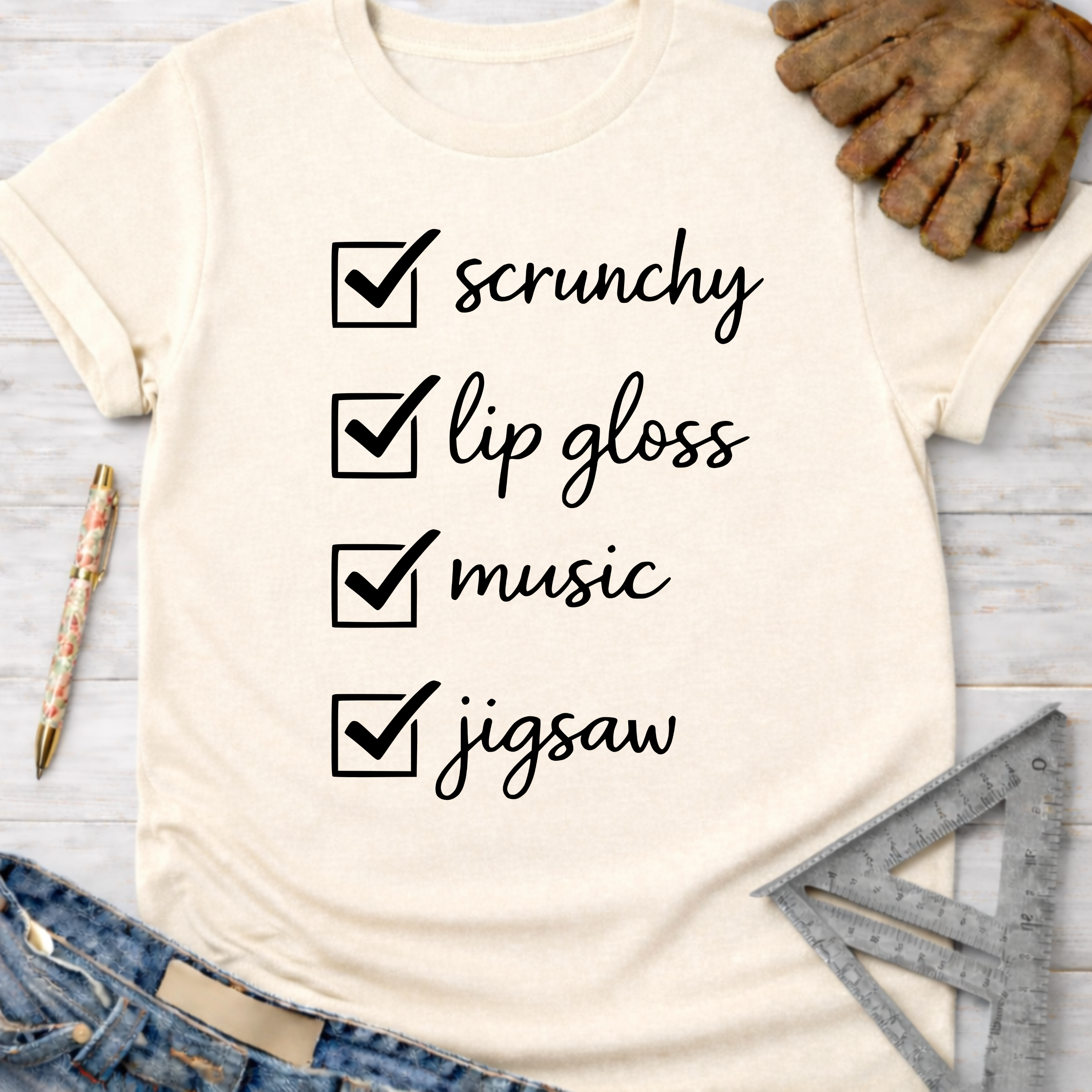 Women's funny woodworking graphic tee shirt for female woodworkers, makers, and DIY builders — Sawdust & Ponytails. Cream t-shirt with "scrunchy, lip gloss, music, jigsaw" checklist design.
