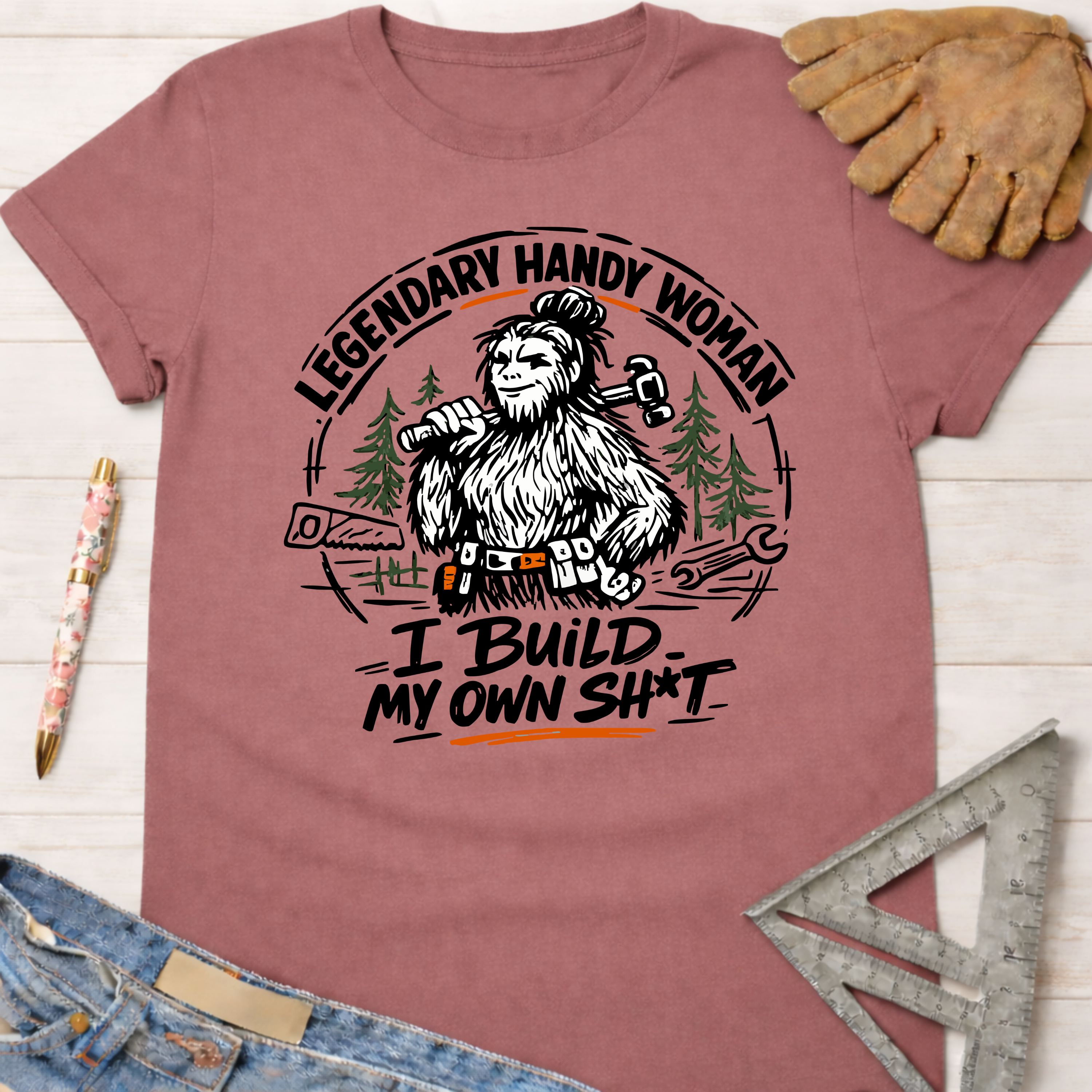Women's funny woodworking graphic tee shirt for female woodworkers, makers, and DIY builders — Sawdust & Ponytails. Mauve t-shirt with a Bigfoot holding a hammer and saw, text "Legendary Handy Woman I Build My Own Sh*t".