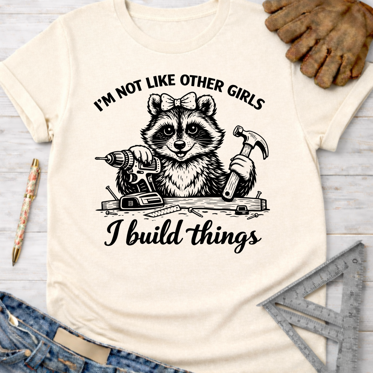 Women's funny woodworking graphic tee shirt for female woodworkers, makers, and DIY builders — Sawdust & Ponytails. Cream t-shirt with a raccoon holding a drill and hammer, text "I'm not like other girls, I build things.