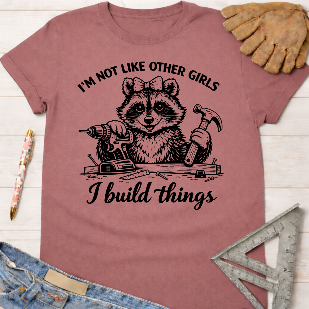 Women's funny woodworking graphic tee shirt for female woodworkers, makers, and DIY builders — Sawdust & Ponytails. Mauve cotton tee with a raccoon holding a drill and hammer, text "I'm not like other girls, I build things.