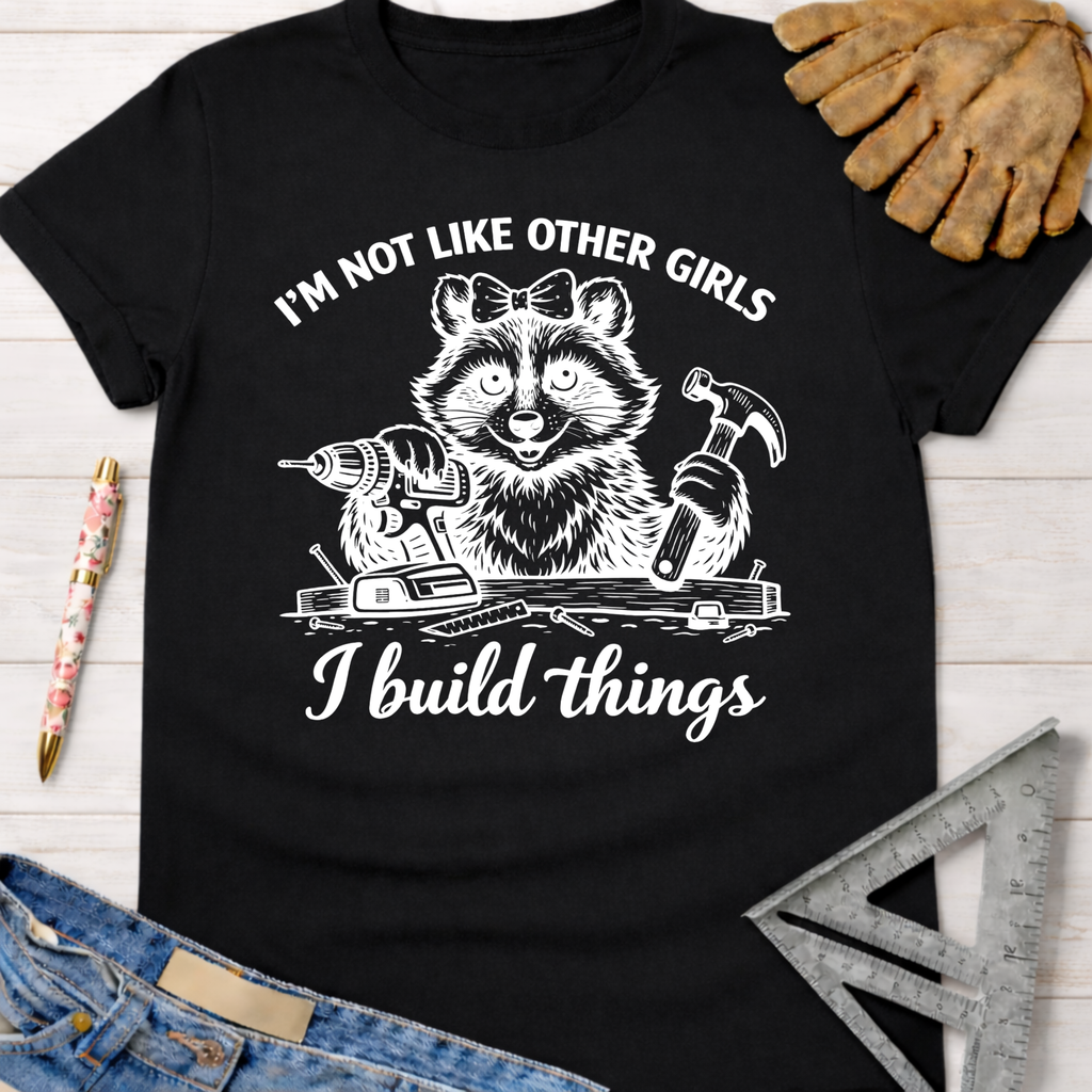 Black cotton women's t-shirt with "I'm Not Like Other Girls, I Build Things" raccoon graphic.