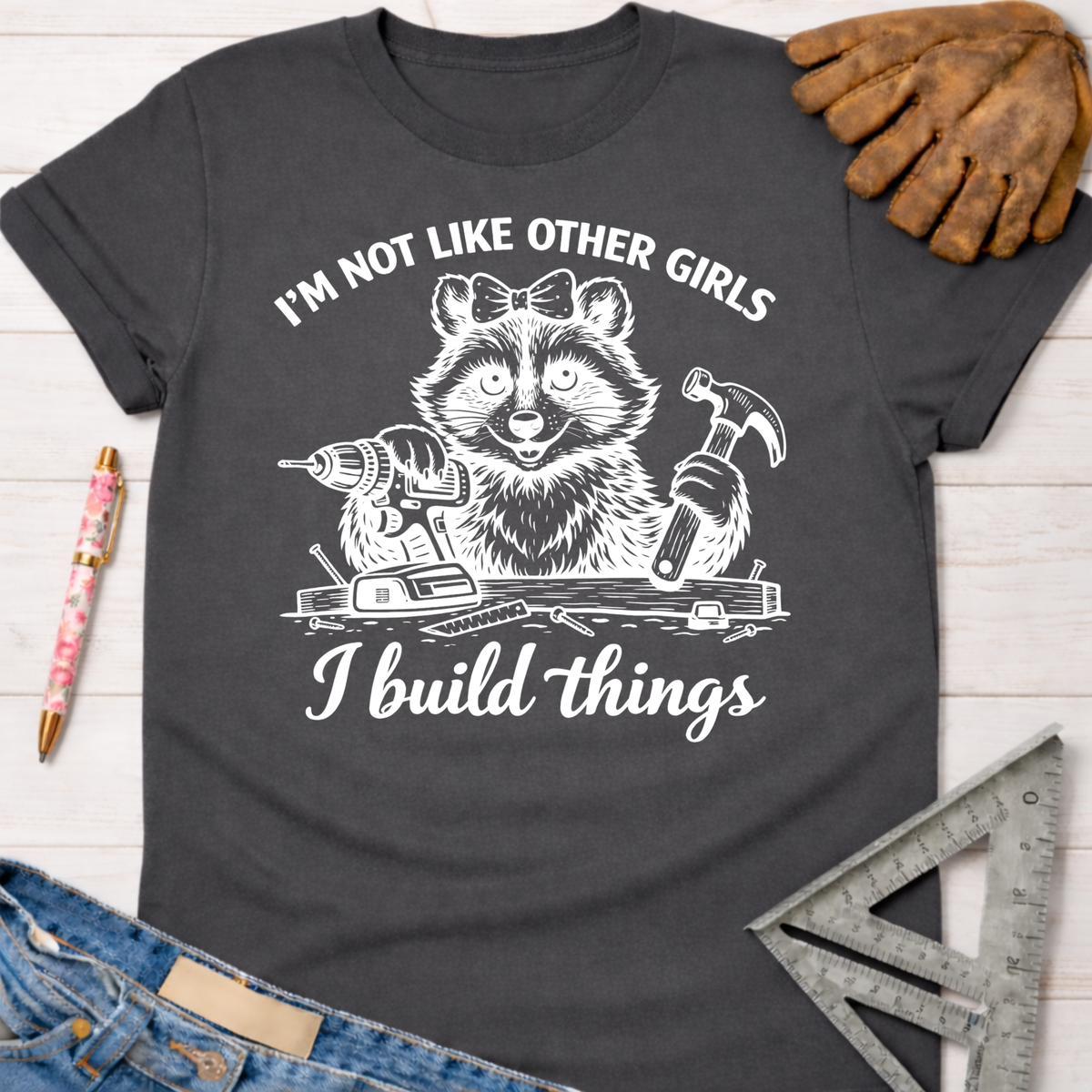 Charcoal gray women's funny woodworking graphic tee with a raccoon holding a drill and hammer, "I build things" text.