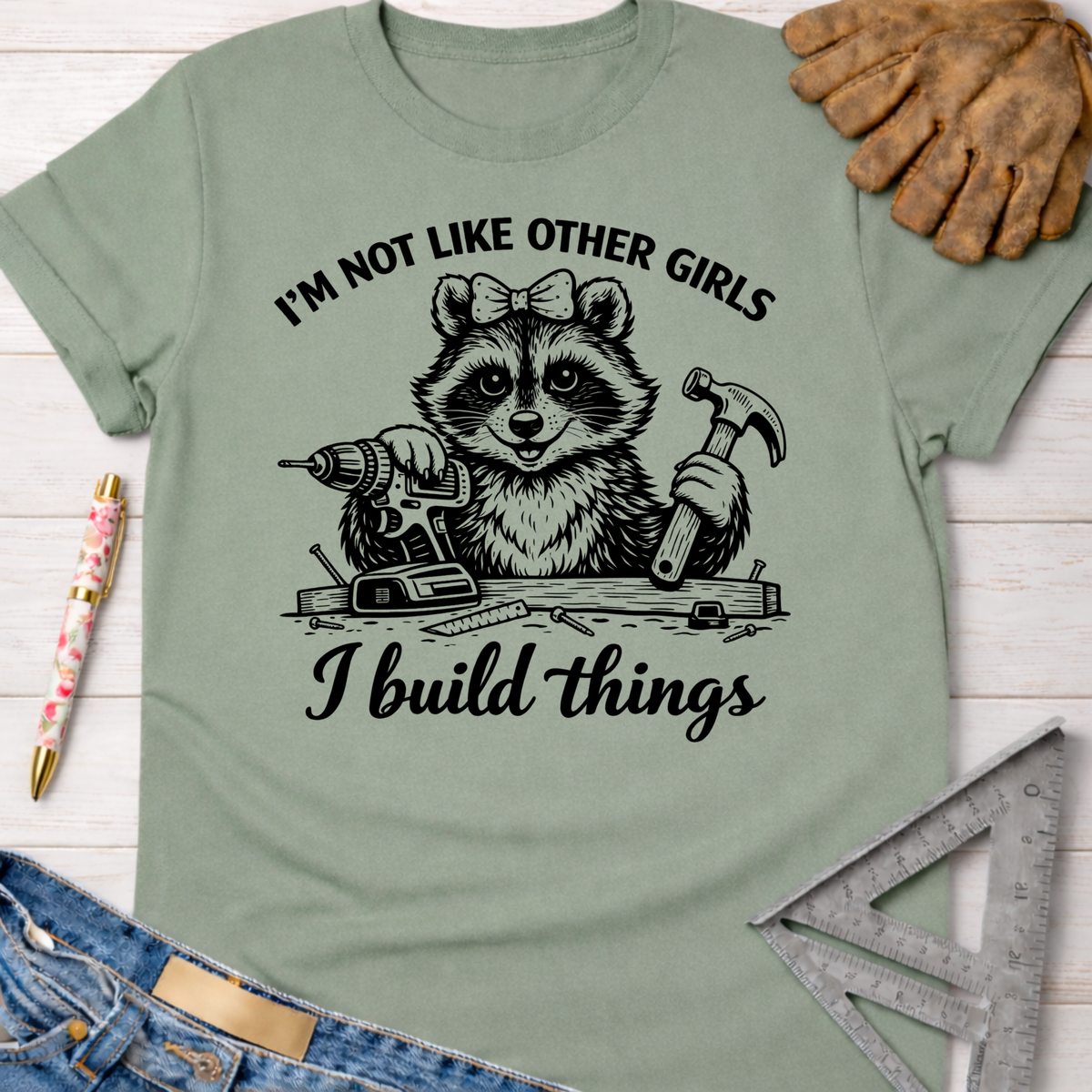 Women's funny woodworking graphic tee shirt for female woodworkers, makers, and DIY builders — Sawdust & Ponytails. Light green cotton tee with a raccoon holding a drill and hammer, text "I'm not like other girls, I build things.