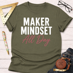 Olive green cotton t-shirt with "MAKER MINDSET All Day" in white and pink text, flat lay.