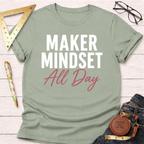 Sage green "Maker Mindset All Day" t-shirt with rolled sleeves, flat laid on a white wooden surface.