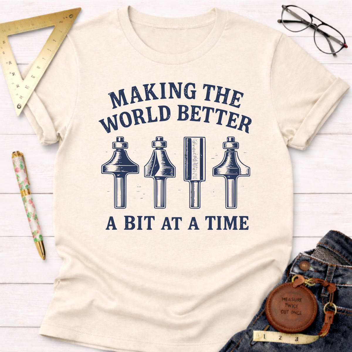 Women's funny woodworking graphic tee shirt for female woodworkers, makers, and DIY builders — Sawdust & Ponytails. Cream cotton t-shirt with "Making The World Better A Bit At A Time" text and router bit graphics.