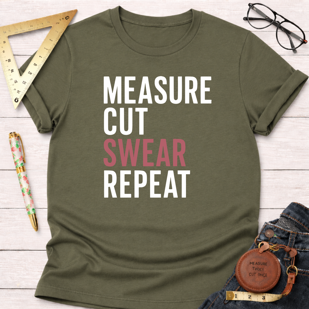 Olive green t-shirt with "MEASURE CUT SWEAR REPEAT" in white and pink text, styled with jeans and tools.