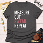 Dark gray "Measure Cut Swear Repeat" graphic tee with rolled sleeves, beside jeans, glasses, and tools.