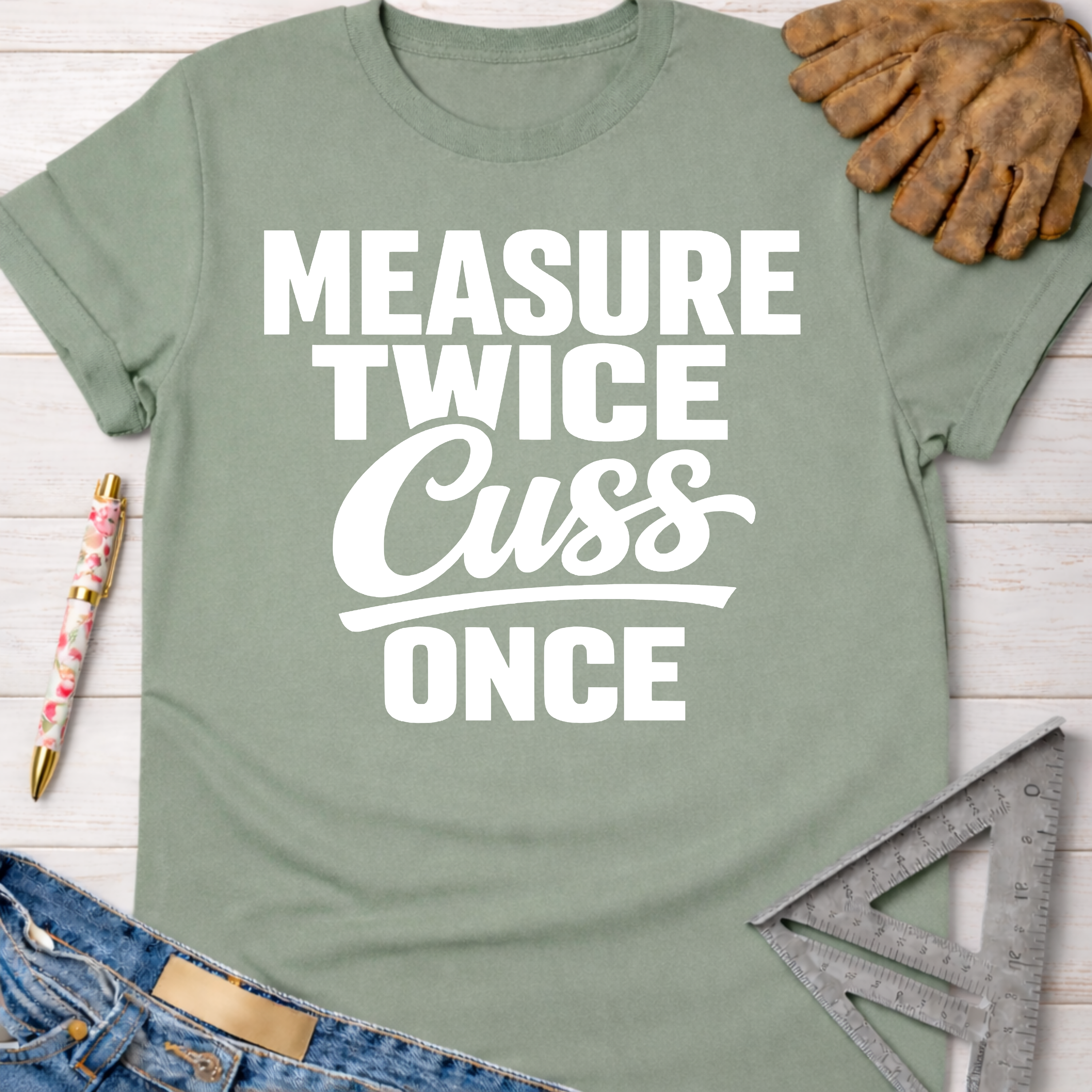 Women's funny woodworking graphic tee shirt for female woodworkers, makers, and DIY builders — Sawdust & Ponytails. Sage green t-shirt with "Measure Twice Cuss Once" in white text.