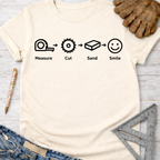 Women's funny woodworking graphic tee shirt for female woodworkers, makers, and DIY builders — Sawdust & Ponytails. Light beige t-shirt with "Measure. Cut. Sand. Smile." icons.